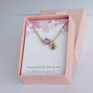 May include: A gold necklace with a purple flower charm and a small gold disc with the letter "H" engraved on it. The necklace is in a pink box with the words "Thank you for being our Flower girl" printed on it.