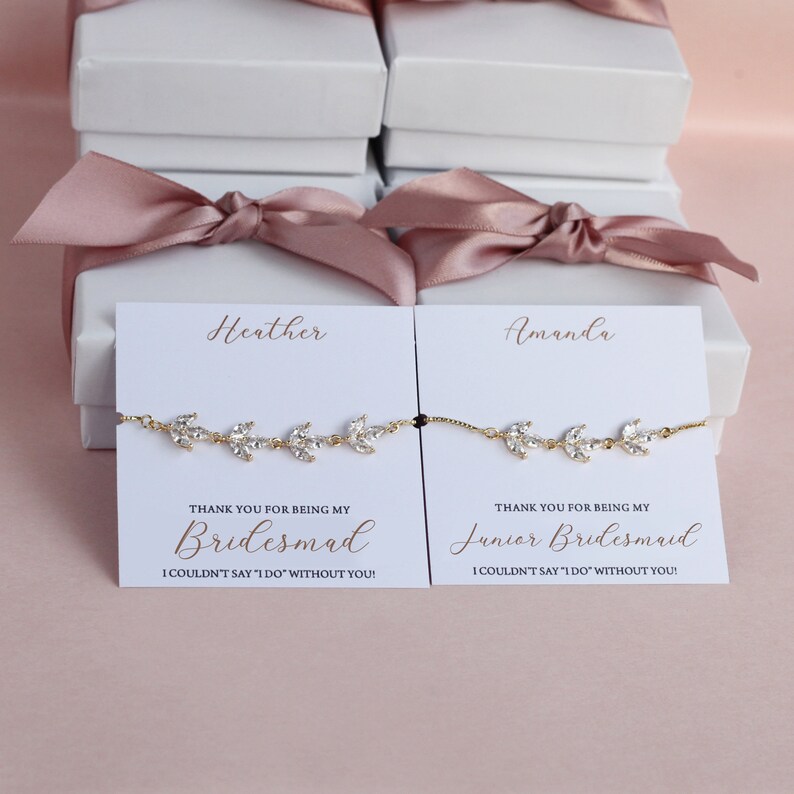 May include: Two white gift boxes with pink ribbon bows, each containing a delicate gold bracelet with clear crystals and a card. The card for the left box reads "Heather" and "Thank you for being my Bridesmaid I couldn't say 'I do' without you!" The card for the right box reads "Amanda" and "Thank you for being my Junior Bridesmaid I couldn't say 'I do' without you!"