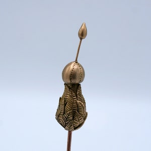 Könnte beinhalten: Eine goldfarbene Metallskulptur einer stilisierten Blume mit einer spitzen Spitze und einer runden Basis. Die Blume besteht aus vielen kleinen, überlappenden Blättern.