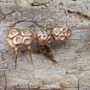 May include: A collection of eight small, gold-colored, ant-shaped sculptures with segmented bodies and six legs. The sculptures are arranged in a row on a piece of weathered wood.