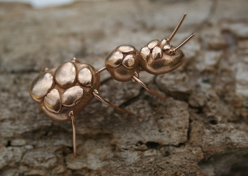 May include: A close-up of a copper ant sculpture. The ant is made of small, rounded pieces of copper that are joined together. The ant has six legs and two antennae. The ant is standing on a piece of wood.