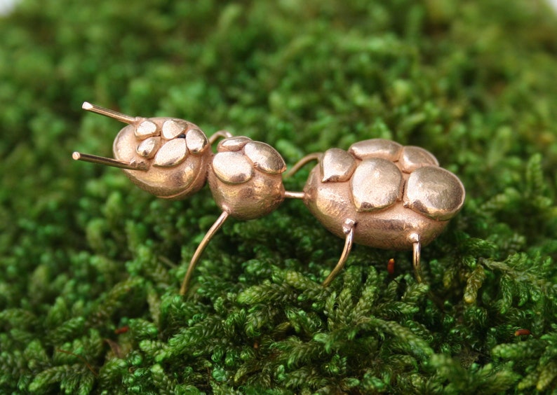 May include: A bronze ant sculpture with a segmented body and six legs. The ant is standing on a bed of green moss.