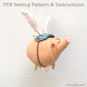 May include: A pink felt pig with white felt wings, wearing a blue and white floral saddle, flying against a white wall. The pig has a brown felt strap around its body and black eyes.