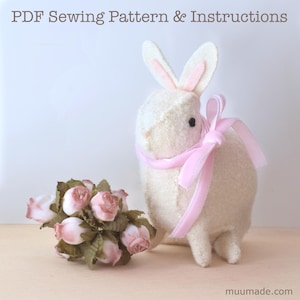 A white felt bunny with a light pink ribbon around its neck and a bouquet of light pink flowers next to it. Its sewing pattern & instructions are available for instant download from muumade.etsy.com.