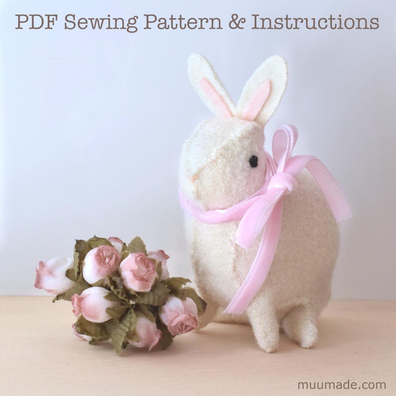 A white felt bunny with a light pink ribbon around its neck and a bouquet of light pink flowers next to it. Its sewing pattern & instructions are available for instant download from muumade.etsy.com.