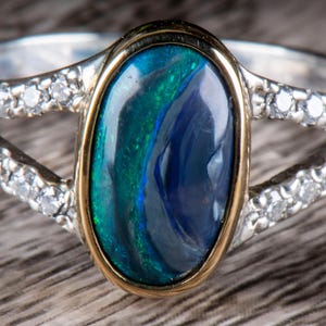 May include: An oval-shaped opal ring with a gold bezel and a silver band. The opal features a vibrant display of blue and green hues. The band is split and adorned with small, sparkling gemstones.
