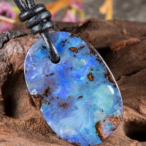 Op de afbeelding: Een ovaalvormige boulder opaal hanger met levendige blauwe en turquoise flitsen en donkerbruine ijzersteen matrix insluitingen. De gepolijste steen hangt aan een zwart leren koord dat door een geboord gat aan de bovenkant is gehaald.