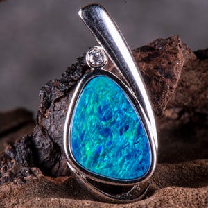 Könnte beinhalten: Ein silberner Anhänger mit einem blauen Opal-Edelstein und einem kleinen klaren Diamanten als Akzent. Der Anhänger ist vor einem dunkelbraunen Holzhintergrund platziert.