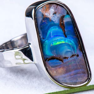 May include: A silver ring featuring a large, rectangular opal. The opal showcases vivid blues, greens, and browns. The ring has a hammered texture on the band and a polished silver bezel setting.