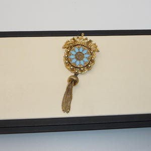 Peut inclure: Broche dorée avec un motif floral bleu clair au centre. La broche présente des détails complexes et une gland de chaînes dorées. Le design rappelle les bijoux vintage.