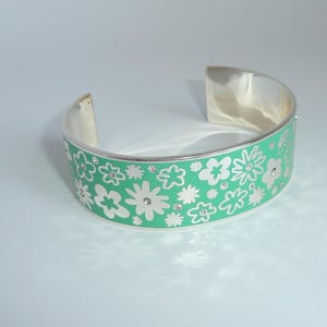 May include: Silver-tone cuff bracelet with a light green enamel band. The band features a floral pattern in white and is accented with small, clear crystals. The bracelet has a modern, open design.