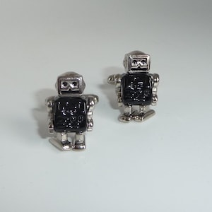 May include: A pair of robot-shaped cufflinks. The cufflinks have a silver-colored metal finish with black enamel detailing on the robot's body. The robot's head and limbs are silver, and the body is black. The cufflinks are designed to be worn on a shirt cuff.
