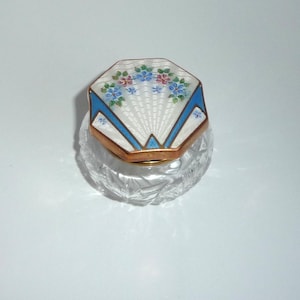 Puede incluir: Un pequeño frasco de vidrio con una base de cristal transparente facetado y una tapa octogonal decorativa. La tapa presenta esmalte guilloché blanco con detalles florales azules y rojos, enmarcado por bordes de esmalte azul brillante y un borde dorado.