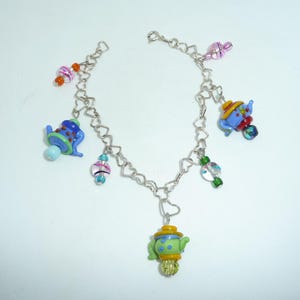 May include: A silver charm bracelet with heart-shaped links and colorful glass teapot charms. The charms are decorated with blue, green, yellow, red, and white paint.