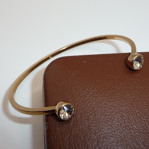 May include: A gold-coloured cuff bracelet with a simple design. The open bracelet has a curved band and is decorated with a clear, faceted gemstone at each end. The bracelet rests on a brown surface.
