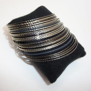 Puede incluir: Un conjunto de pulseras de color plateado con una superficie texturizada, apoyadas sobre un pequeño cojín negro. Las pulseras son finas y parecen ser de metal. El fondo es blanco.