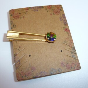 Puede incluir: Un pasador de corbata de metal dorado con un diseño de marco rectangular abierto y alargado. Un extremo del pasador está adornado con una piedra preciosa redonda e iridiscente en un engaste de cuatro puntas, que muestra tonos verdes y morados.