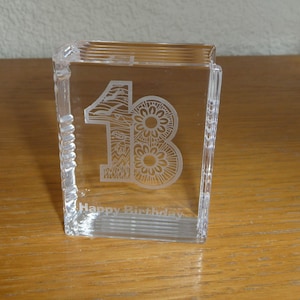 May include: A clear glass paperweight with the number 18 etched in a decorative design. The paperweight also has the words "Happy Birthday" etched on the bottom.