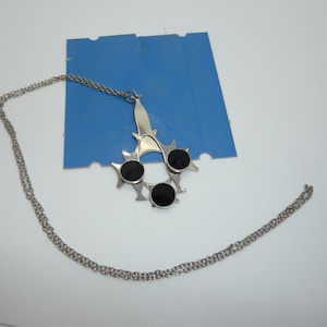 Può includere: Una collana d'argento con un ciondolo con un design stilizzato con tre pietre nere. Il ciondolo è su uno sfondo blu.