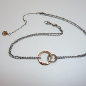 Puede incluir: Un collar de doble cadena plateada con dos anillos entrelazados como colgante central. Un anillo es de tono dorado con texto grabado, mientras que el otro es plateado. El collar incluye un cierre de mosquetón y un pequeño dije circular.
