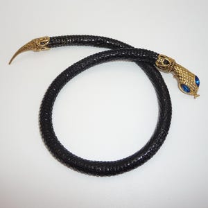 May include: A flexible black snake-shaped necklace with a textured scale pattern. The piece features a gold-toned metal head with two bright blue inset eyes and a matching gold-toned pointed tail end, creating a continuous serpent-like design.