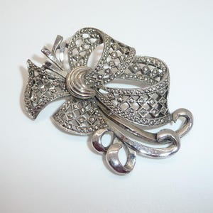 May include: A silver-toned brooch in the shape of a bow with a delicate, intricate design. The bow is made of a filigree pattern with small, round stones.