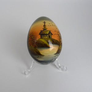 May include: A decorative egg featuring a hand-painted landscape with a small wooden church on a hill overlooking a body of water. The scene is set against an orange and yellow sunset sky. The egg rests on a clear, three-legged acrylic stand.