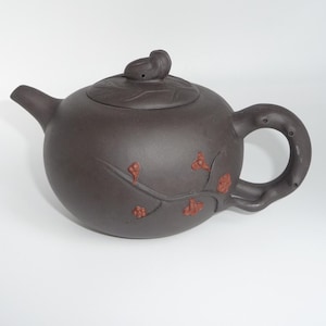 May include: A dark brown clay teapot featuring a rounded body and a lid topped with a small sculpted snail. The exterior is decorated with embossed reddish-brown floral branch motifs, and the handle is shaped to resemble a textured tree branch.