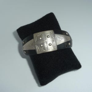 Puede incluir: Una pulsera de brazalete ancha de color negro con una placa central rectangular de metal plateado que presenta un patrón geométrico grabado y cuatro pequeños remaches circulares. La banda negra tiene detalles de incrustaciones de metal plateado.