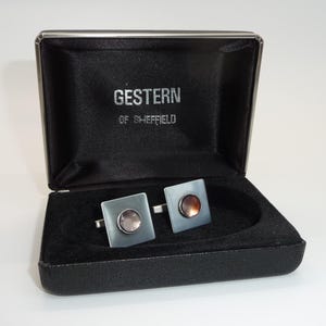 Può includere: Una scatola nera aperta con un paio di gemelli quadrati. I gemelli hanno una base quadrata grigio argento con un centro circolare color rame. Il testo "GESTERN OF SHEFFIELD" è visibile all'interno della scatola.