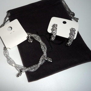 May include: A silver-toned mesh bracelet featuring small dangling heart charms and a pair of matching hoop earrings with heart-shaped details. The jewellery pieces are displayed on white cardboard tags resting atop a dark brown velvet drawstring pouch.