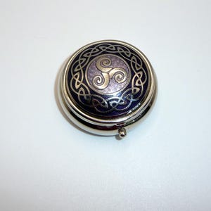 May include: A round, silver-toned metal pill box featuring a deep purple enamel inlay on the lid. The lid is decorated with a silver-coloured triskelion symbol at the centre, surrounded by an intricate, continuous silver Celtic knotwork pattern.