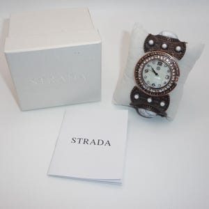May include: A Strada watch with a white face, surrounded by a ring of crystals, and a bronze-colored band. The watch rests on a white pillow. A white box and a booklet with the word "STRADA" are also in the image.