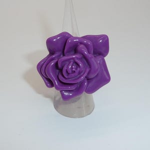 May include: A large, glossy, purple rose ring. The ring features a detailed floral design with multiple petals and a smooth, shiny finish. The ring is displayed on a clear, cone-shaped stand.