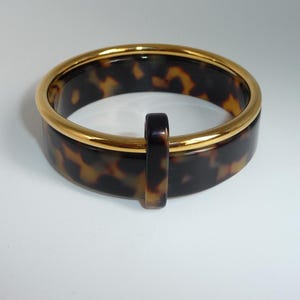 Może przedstawiać: Okrągła bransoletka typu bangle z brązowo-czarnym wzorem szylkretowym. Bransoletka jest wykończona gładką, polerowaną złotą metalową krawędzią wzdłuż górnego brzegu oraz małą, pionową szylkretową wypustką przymocowaną z boku.