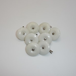 May include: Eight white ceramic cabinet knobs with silver metal fixings. The knobs are round with a slightly domed surface and a small, central screw. They are arranged on a white background, suitable for furniture or decor.