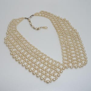 Puede incluir: Un collar de perlas color crema con un diseño calado ancho. El collar presenta una serie de hilos de perlas interconectados, creando una forma de cuello. Tiene un cierre plateado y una cadena ajustable.