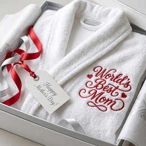 Puede incluir: Una bata blanca de rizo con el texto bordado "World's Best Mom" en rojo, presentada en una caja de regalo. Una etiqueta con "Happy Mother's Day!" está sujeta con cintas rojas y plateadas. La bata está doblada.