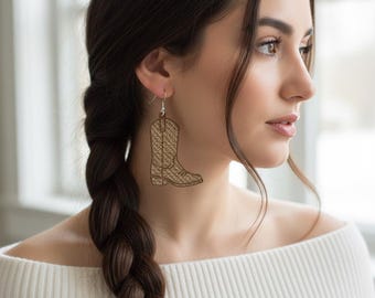 May include: A single earring shaped like a cowboy boot. The boot is a light brown color with a woven texture. The earring hangs from a silver hook. The background is blurred, but appears to be a window.