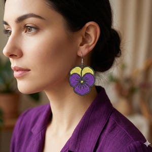Puede incluir: Pendientes de flores de pensamiento bordadas en morado y amarillo, con contorno negro y ganchos plateados. La persona lleva una camisa morada. Un accesorio floral.