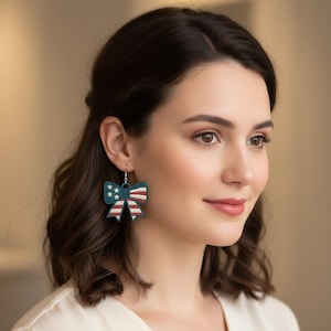 May include: Bow-shaped earrings with a patriotic design. The earrings feature a dark teal background with white stars and red and white stripes, resembling the American flag. The earrings are hanging from a silver hook.