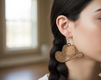 May include: A close-up of a person wearing a cowboy hat earring. The earring is a detailed, laser-cut design of a cowboy hat. The hat is a light brown color with a woven texture, and the earring hangs from a silver hook.