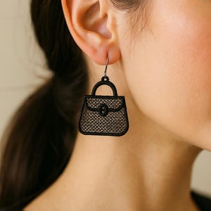 May include: Black handbag-shaped earrings with a textured, open-weave design. The earrings have a small handle and a clasp detail. The earrings are suspended from a silver-colored hook.