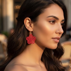 Puede incluir: Pendientes rojos con forma de flor con intrincados detalles de encaje. Los pendientes están suspendidos de ganchos dorados. El diseño presenta una flor de cinco pétalos con un centro detallado, creando un accesorio delicado.