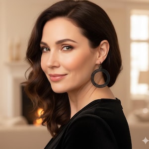 以下が含まれることがあります： 黒いフープイヤリングをつけた、濃い波状の髪の女性。イヤリングはテクスチャード加工のデザインです。彼女は黒いトップスを着て、カメラを見ています。背景はぼやけており、暖炉と窓が見えます。