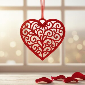 Puede incluir: Un adorno rojo en forma de corazón con un diseño intrincado y arremolinado cuelga de una cinta roja. Pétalos de rosa rojos están esparcidos sobre una superficie de madera frente a una ventana con iluminación suave y borrosa.