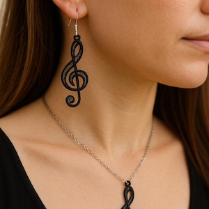 Puede incluir: Juego de pendientes y collar con clave de sol negra. Los pendientes cuelgan de ganchos plateados, y el collar presenta un colgante a juego en una cadena plateada. La joyería de temática musical es un accesorio elegante.