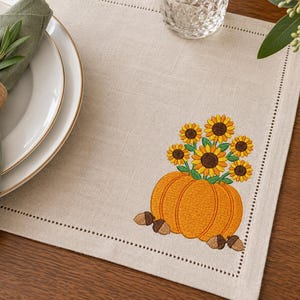 May include: A square beige placemat featuring an embroidered design of a large orange pumpkin topped with a bouquet of yellow sunflowers and green leaves, accented by small brown acorns at the base, with a decorative stitched border detail.