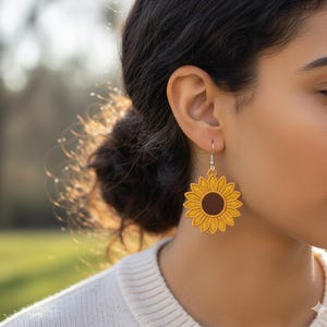 Può includere: Primo piano di una persona che indossa orecchini a forma di girasole. Gli orecchini sono gialli con un centro marrone e hanno la forma di girasoli. Gli orecchini sono appesi a ganci d'argento.