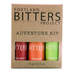 May include: A cardboard box with a window showing three small bottles of bitters. The bottles are labeled "Pepper Spice", "Orange", and "Woodlan" and are part of the "Portland Bitters Project" Adventure Kit. The box is labeled "3 x 0.5 fl oz".